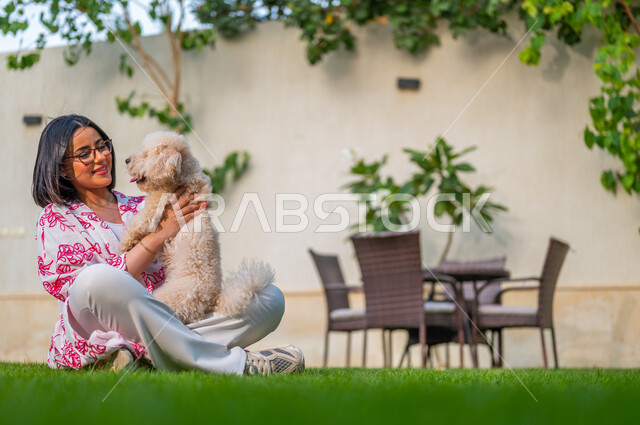 اللعب والمرح مع الحيوانات الأليفة، مفهوم الرفق بالحيوان، تربية واقتناء الكلاب المنزلية، امرأة عربية خليجية سعودية تجلس على العشب وتحمل جرو صغير، الشعور بالسعادة أثناء قضاء وقت ممتع في أيام العطلة