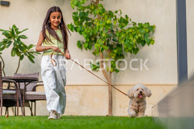 الشعور بالسعادة أثناء قضاء وقت ممتع في أيام العطلة، اللعب والمرح مع الحيوانات الأليفة، مفهوم الرفق بالحيوان، تربية واقتناء الكلاب المنزلية، فتاة عربية خليجية سعودية تمشي على العشب ومعها جرو صغير