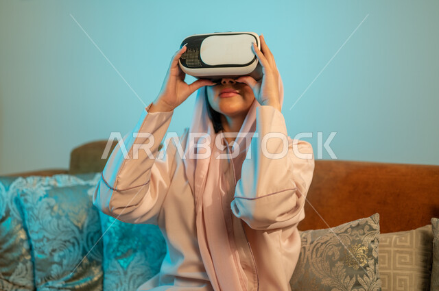 الاستمتاع بمشاهدة أفلام العالم الخيالي، استخدام الأجهزة والتقنيات الحديثة، قضاء اوقات ممتعة في المنزل، إيماءات الاندماج والتفاعل مع الألعاب الافتراضية،  إمرأة عربية خليجية سعودية ترتدي الحجاب والعباءة تضع نظارة الواقع الافتراضي ثلاثية الابعاد
