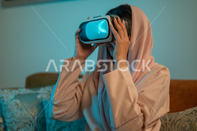 قضاء اوقات ممتعة في المنزل، استخدام الأجهزة والتقنيات الحديثة، إيماءات الاندماج والتفاعل مع الألعاب الافتراضية، الاستمتاع بمشاهدة أفلام العالم الخيالي، إمرأة عربية خليجية سعودية ترتدي الحجاب والعباءة تضع نظارة الواقع الافتراضي ثلاثية الابعاد