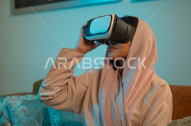 إيماءات الاندماج والتفاعل مع الألعاب الافتراضية، الاستمتاع بمشاهدة أفلام العالم الخيالي، إمرأة عربية خليجية سعودية ترتدي الحجاب والعباءة تضع نظارة الواقع الافتراضي ثلاثية الابعاد، قضاء اوقات ممتعة في المنزل، استخدام الأجهزة والتقنيات الحديثة