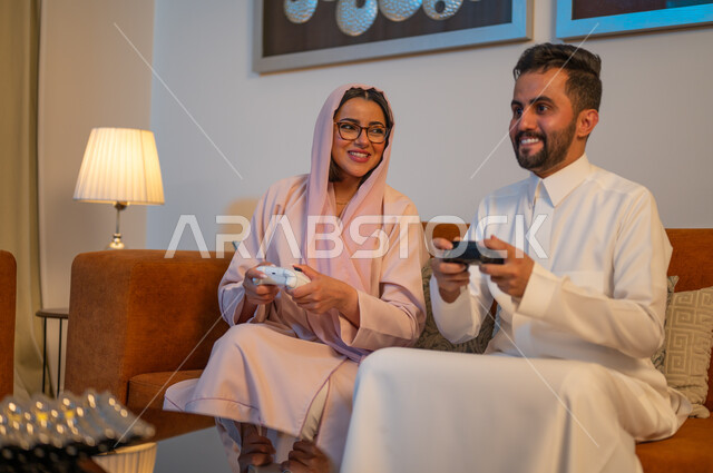 الاندماج والتفاعل مع ألعاب الفيديو، قضاء وقت ممتع ومسلي في المنزل، زوجان عربيان خليجيان سعوديان يمسكان عصا التحكم بتعابير السرور والسعادة، فعاليات عطلة نهاية الأسبوع، تقنيات الكترونية حديثة ترفيهيه