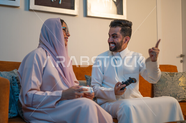 قضاء وقت ممتع ومسلي في المنزل، التفاعل والاندماج مع ألعاب الفيديو، زوجان عربيان خليجيان سعوديان يمسكان عصا التحكم بتعابير السرور والسعادة، فعاليات عطلة نهاية الأسبوع، تقنيات الكترونية حديثة ترفيهيه