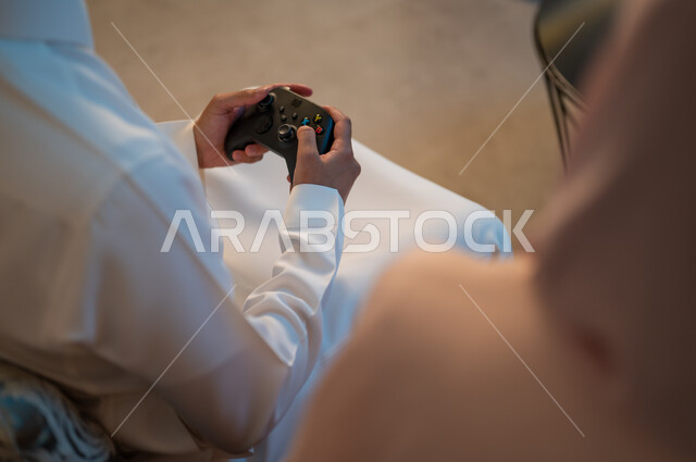 فعاليات عطلة نهاية الأسبوع، تقنيات الكترونية حديثة ترفيهيه, قضاء وقت ممتع ومسلي في المنزل، التفاعل والاندماج مع ألعاب الفيديو، زوجان عربيان خليجيان سعوديان يمسكان عصا التحكم بتعابير السرور والسعادة