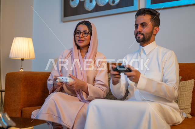 Expressions of enthusiasm, integration, and interaction with video games, spending a fun and entertaining time at home, a Saudi Gulf Arab couple holding the joystick with gestures of pleasure and happiness, weekend activities, modern electronic entertainment technologies