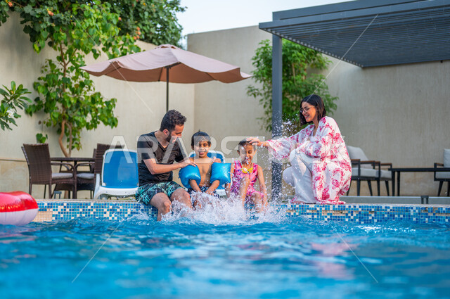 The concept of a happy, interconnected family, fun recreational trips, spending enjoyable and refreshing times in the summer, enjoying one of the Kingdom’s swimming pools, sitting on the edge of the water pool, a recreational tourist trip, a smiling Saudi Gulf Arab family wearing a swimsuit.