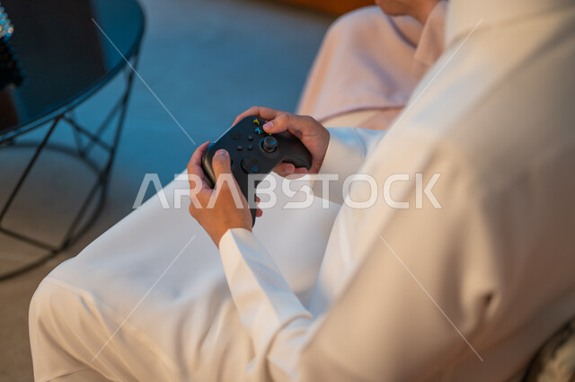 قضاء وقت ممتع ومسلي في المنزل، التفاعل والاندماج مع ألعاب الفيديو، زوجان عربيان خليجيان سعوديان يمسكان عصا التحكم بتعابير السرور والسعادة، فعاليات عطلة نهاية الأسبوع، تقنيات الكترونية حديثة ترفيهيه