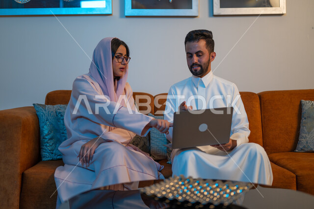 استخدام الأجهزة والتقنيات الحديثة والمتطورة، رجل عربي خليجي سعودي يقوم باستخدام الحاسوب المحمول بجانب زوجته، إنجاز المهام في المنزل، التعاون وتبادل الآراء والمشاركة بين الزوجين، ادارة الأعمال عن بعد