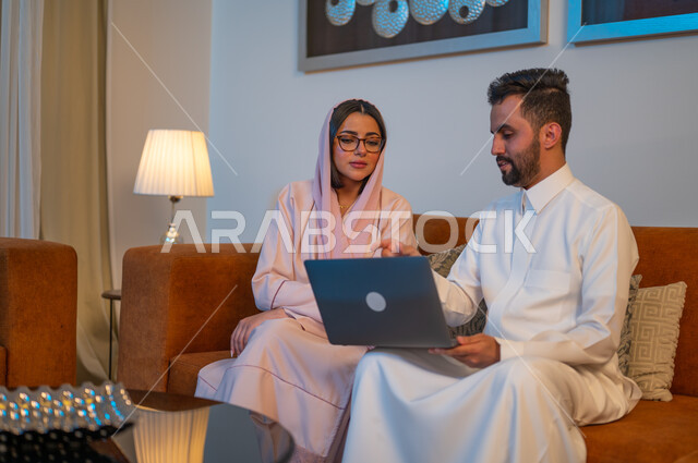 إنجاز المهام في المنزل، التعاون وتبادل الآراء والمشاركة بين الزوجين، ادارة الأعمال عن بعد، استخدام الأجهزة والتقنيات الحديثة والمتطورة، رجل عربي خليجي سعودي يقوم باستخدام الحاسوب المحمول بجانب زوجته