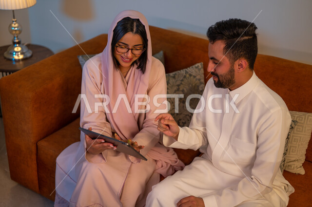 ادارة الأعمال عن بعد، استخدام الأجهزة والتقنيات الحديثة والمتطورة، إنجاز المهام في المنزل، المشاركة والتعاون وتبادل الآراء بين الزوجين، إمرأة عربية خليجية سعودية ترتدي العباءة والحجاب تقوم باستخدام جهاز التابليت اللوحي بجانب زوجها