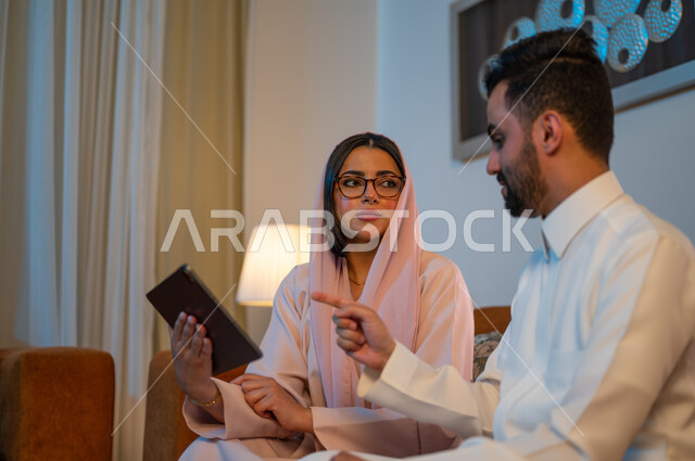 إنجاز المهام في المنزل، المشاركة والتعاون وتبادل الآراء بين الزوجين، ادارة الأعمال عن بعد، استخدام الأجهزة والتقنيات الحديثة والمتطورة، إمرأة عربية خليجية سعودية ترتدي العباءة والحجاب تقوم باستخدام جهاز التابليت اللوحي بجانب زوجها