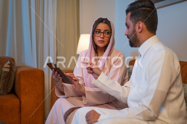 مفهوم إدارة شؤون الأعمال وتنظيمها عبر الاجهزة الحديثة والمتطورة، العمل عن بعد على مواقع التواصل الاجتماعي، المشاركة والتعاون وتبادل الآراء بين الزوجين، إمرأة عربية خليجية سعودية ترتدي العباءة والحجاب تقوم باستخدام جهاز التابليت اللوحي بجانب زوجها