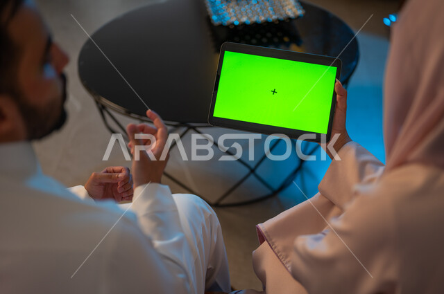 كروما، إدارة شؤون الأعمال وتنظيمها عبر الاجهزة الحديثة والمتطورة، العمل عن بعد على مواقع التواصل الاجتماعي، المشاركة والتعاون وتبادل الآراء بين الزوجين، إمرأة عربية خليجية سعودية ترتدي العباءة تقوم باستخدام جهاز التابليت اللوحي بشاشة خضراء فارغة بجانب زوجها