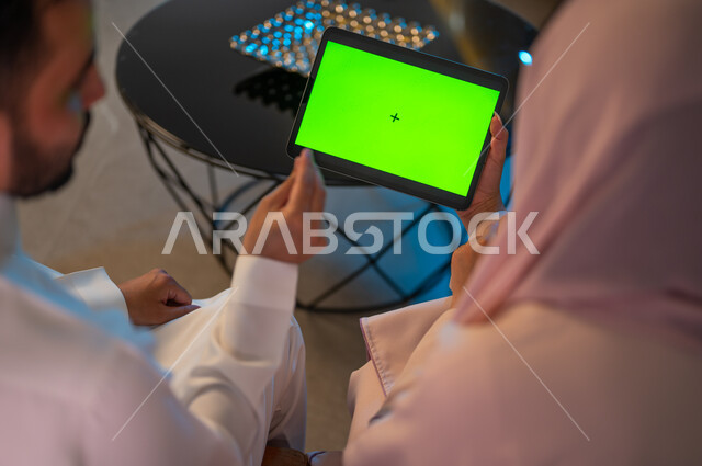 كروما، إدارة شؤون الأعمال وتنظيمها عبر الاجهزة الحديثة والمتطورة، العمل عن بعد على مواقع التواصل الاجتماعي، المشاركة والتعاون وتبادل الآراء بين الزوجين، إمرأة عربية خليجية سعودية ترتدي العباءة تقوم باستخدام جهاز التابليت اللوحي بشاشة خضراء فارغة بجانب زوجها