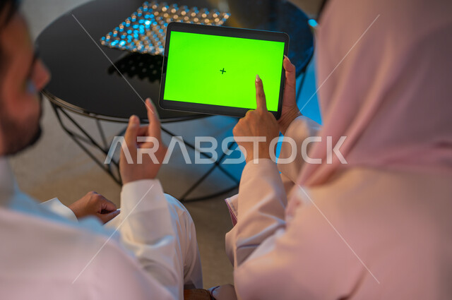 كروما، إدارة شؤون الأعمال وتنظيمها عبر الاجهزة الحديثة والمتطورة، العمل عن بعد على مواقع التواصل الاجتماعي، المشاركة والتعاون وتبادل الآراء بين الزوجين، إمرأة عربية خليجية سعودية ترتدي العباءة تقوم باستخدام جهاز التابليت اللوحي بشاشة خضراء فارغة بجانب زوجها