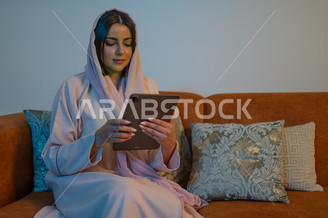 إنجاز المهام العملية عبر جهاز التابليت، التسوق الالكتروني عبر الأونلاين، استخدام الأجهزة والتقنيات الحديثة والمتطورة، إمرأة عربية خليجية سعودية ترتدي العباءة والحجاب تقوم باستخدام الجهاز اللوحي، إنجاز المهام في المنزل، مهن ووظائف سعودية نسائية