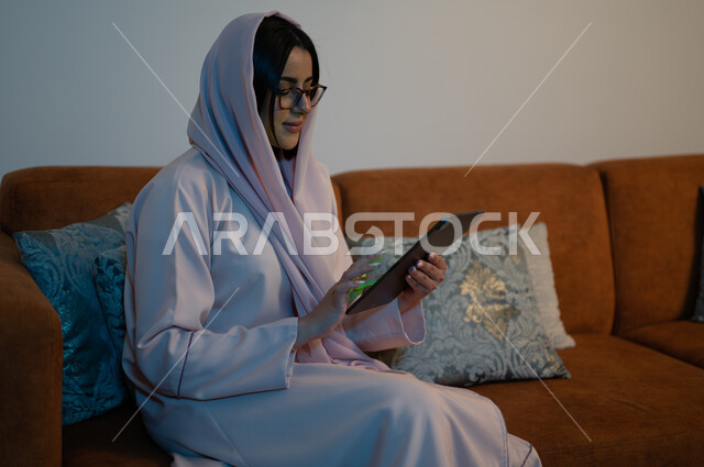 مهن ووظائف سعودية نسائية، إنجاز المهام العملية عبر جهاز التابليت، التسوق الالكتروني عبر الأونلاين، استخدام الأجهزة والتقنيات الحديثة والمتطورة، إمرأة عربية خليجية سعودية ترتدي العباءة والحجاب تقوم باستخدام الجهاز اللوحي، إنجاز المهام في المنزل