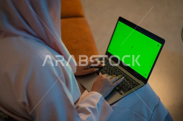 دمج التكنولوجيا والتقنيات لمتابعة وتطوير الأعمال، تصوير مقرب من الخلف لإمرأة عربية خليجية سعودية تقوم بالكتابة على لوحة مفاتيح جهاز الحاسوب المحمول بشاشة خضراء فارغة، إنجاز المهام في المنزل، مهن ووظائف سعودية نسائية، استخدام الأجهزة والتقنيات الحديثة، كروما