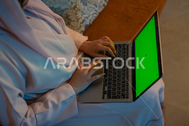 Using modern devices and techniques, integrating technology and techniques to follow up and develop business, close-up photography from the back of a Saudi Gulf Arab woman typing on a laptop keyboard with a blank green screen, completing tasks at home, Saudi women’s professions and jobs, chroma
