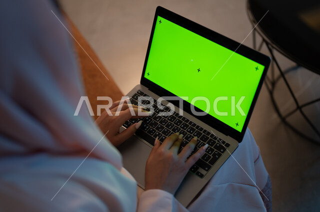Using modern devices and techniques, integrating technology and techniques to follow up and develop business, close-up photography from the back of a Saudi Gulf Arab woman typing on a laptop keyboard with a blank green screen, completing tasks at home, Saudi women’s professions and jobs, chroma