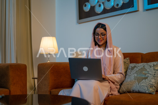 مهن ووظائف سعودية نسائية، ادارة الأعمال عن بعد، استخدام الأجهزة والتقنيات الحديثة والمتطورة، إمرأة عربية خليجية سعودية ترتدي العباءة والحجاب تقوم باستخدام الحاسب المحمول، إنجاز المهام في المنزل، تعابير وايماءات التركيز