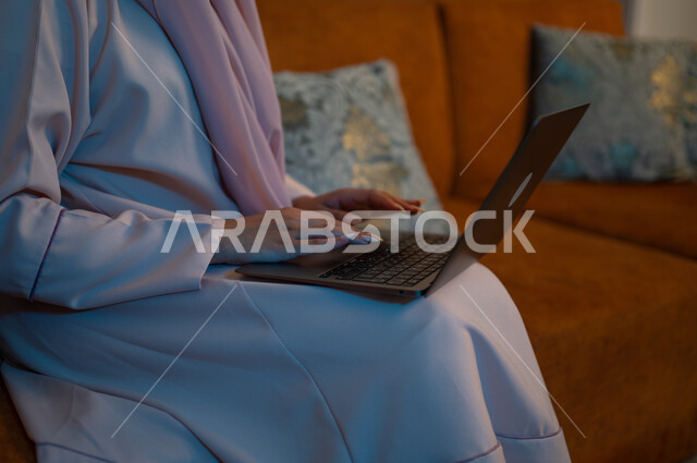 دمج التكنولوجيا بالعمل، مهن ووظائف سعودية نسائية، ادارة الأعمال عن بعد، استخدام الأجهزة والتقنيات الحديثة والمتطورة، إمرأة عربية خليجية سعودية ترتدي العباءة والحجاب تقوم باستخدام الحاسب المحمول، إنجاز المهام في المنزل، تعابير وايماءات التركيز