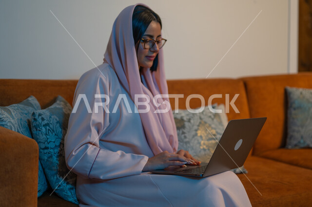 مفهوم دمج التكنولوجيا بالعمل، مهن ووظائف سعودية نسائية، ادارة الأعمال عن بعد، استخدام الأجهزة والتقنيات الحديثة والمتطورة، إمرأة عربية خليجية سعودية ترتدي العباءة والحجاب تقوم باستخدام الحاسب المحمول، إنجاز المهام في المنزل، تعابير وايماءات التركيز