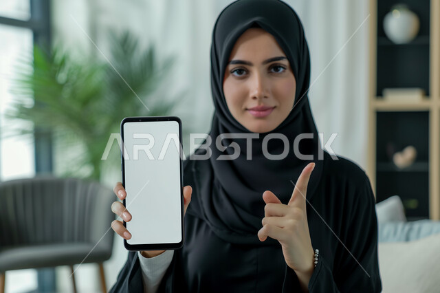 Displaying mobile phone applications and services, mockup on modern technical devices, close-up portrait of a veiled Saudi Gulf Arab woman holding a cell phone with a blank white screen in her hand, raising her index finger up and pointing at something
