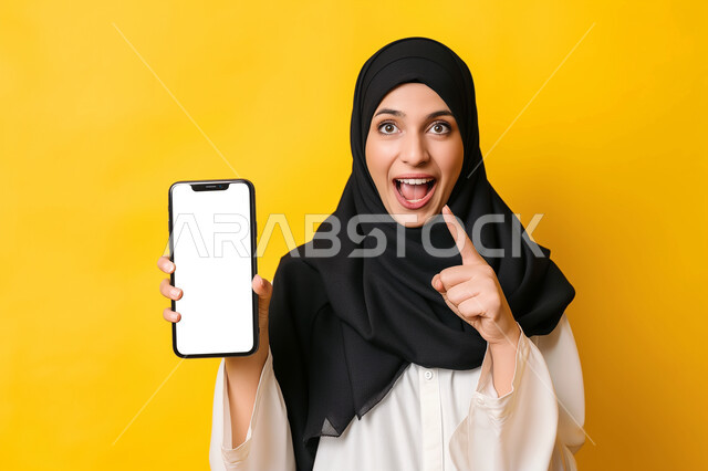 موك أب على الاجهزة التقنية الحديثة، بورتريه مقرب لامرأة عربية خليجية سعودية محجبة تحمل بيدها جوال بشاشة بيضاء فارغه، رفع اصبع السبابة للاعلى و الاشارة الى شيء ما ,إيماءات فم ويدين تدل على المفاجئة والسرور، عرض تطبيقات وخدمات الهاتف المحمول، خلفية صفراء