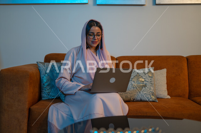 Expressions of focus and integration, integrating technology into work, Saudi women’s professions and jobs, managing business remotely, using modern and advanced devices and technologies, a Saudi Arabian Gulf woman wearing the abaya and hijab, using a laptop, completing tasks at home