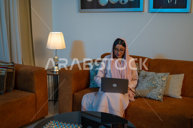 Expressions of focus and integration, integrating technology into work, Saudi women’s professions and jobs, managing business remotely, using modern and advanced devices and technologies, a Saudi Arabian Gulf woman wearing the abaya and hijab, using a laptop, completing tasks at home