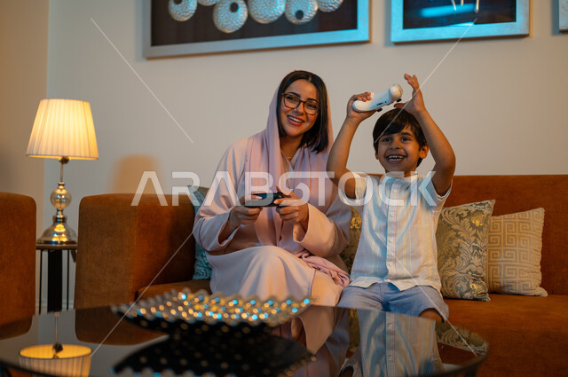 Weekend activities, modern electronic entertainment technologies, spending a fun and entertaining time at home, interacting and merging with video games, a Saudi Arabian Gulf mother wearing the hijab and abaya sitting on the sofa next to her son holding the joystick with expressions of pleasure and happiness.