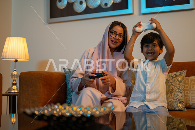 قضاء وقت ممتع ومسلي في المنزل، التفاعل والاندماج مع ألعاب الفيديو، أم عربية خليجية سعودية ترتدي الحجاب والعباءة تجلس على الكنبة بجانب ابنها يمسكان عصا التحكم بتعابير السرور والسعادة، فعاليات عطلة نهاية الأسبوع، تقنيات الكترونية حديثة ترفيهيه
