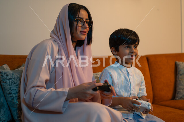 تقنيات الكترونية حديثة ترفيهيه، قضاء وقت ممتع ومسلي في المنزل، التفاعل والاندماج مع ألعاب الفيديو، أم عربية خليجية سعودية ترتدي الحجاب والعباءة تجلس على الكنبة بجانب ابنها يمسكان عصا التحكم بتعابير السرور والسعادة، فعاليات عطلة نهاية الأسبوع