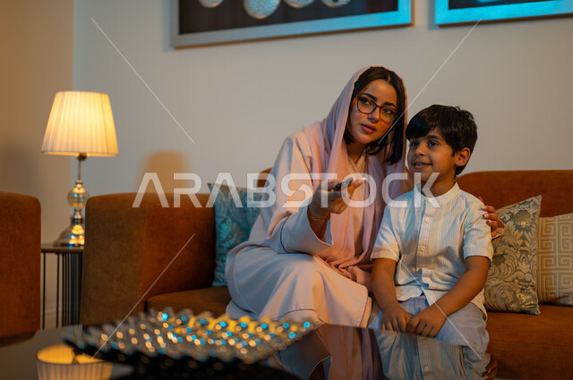 ايماءات الاندماج والتركيز، أجواء اسرية سعيدة في المنزل، أم عربية خليجية سعودية ترتدي العباءة والحجاب تقوم بمشاهدة التلفاز شرب العصير برفقه ابنها، قضاء اوقات عائلية ممتعة، متابعة الافلام والمسلسلات، فلل و شقق سكنية فاخرة في السعودية