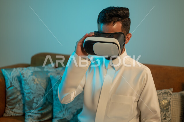 Gestures of integration and interaction with virtual games, enjoying watching movies of the imaginary world, a Saudi Arabian Gulf man wearing a traditional dress, wearing 3D virtual reality glasses, spending enjoyable times at home, using modern devices and technologies.