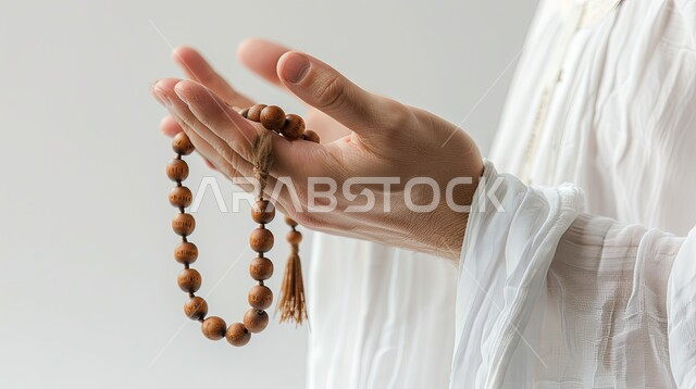 الإكثار من ذكر الله تعالى والإستغفار والدعاء, تصوير مقرب لأيدي رجل عربي سعودي خليجي يرتدي الثوب الأبيض يرفع يديه يمسك السبحه, مفهوم الإجتهاد في العباده و التقرب الى الله في شهر رمضان المبارك, يوم الجمعه المباركه، خلفيه بيضاء