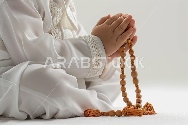 مفهوم الإجتهاد في العباده و التقرب الى الله في شهر رمضان المبارك, الإكثار من ذكر الله تعالى والإستغفار والدعاء, تصوير مقرب لأيدي ولد عربي سعودي خليجي يرتدي الثوب الأبيض يرفع يديه يمسك السبحه, يوم الجمعه المباركه، خلفيه بيضاء