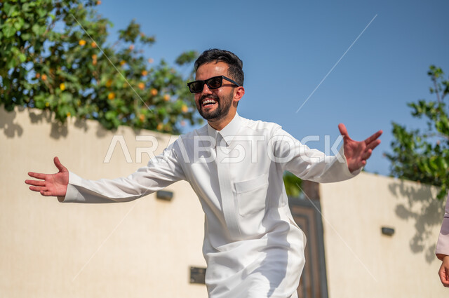 ايماءات تدل على الحماس والسعاده، الإجتماع مع العائله وفرحة العوده من السفر، رجل عربي خليجي سعودي يرتدي الثوب التقليدي يفتح يديه لاحتضان شخص ما