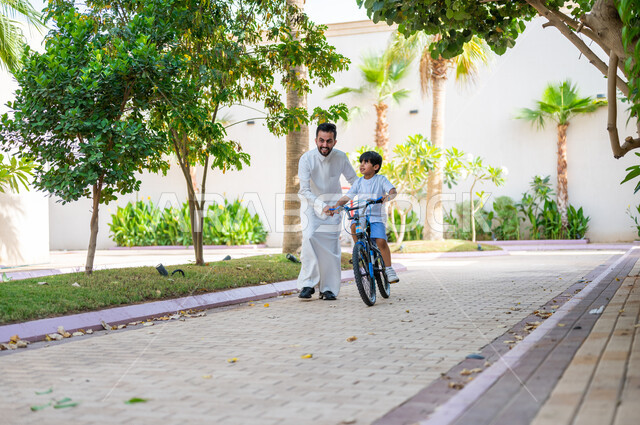 دور الأباء في حياة الأبناء، تعابير الفرح في قضاء أوقات عائلية ممتعة، المشاركة في الأنشطة الترفيهية، مساعدة الأولاد على التوازن واللعب في الدراجات الهوائية، أب عربي خليجي سعودي يرتدي الزي التقليدي يعلم إبنه قيادة الدراجة في إحدى الحدائق العامة