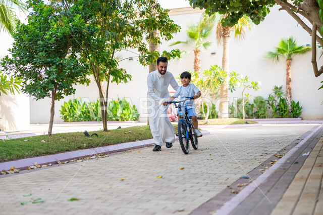 تعابير الفرح في قضاء أوقات عائلية ممتعة، المشاركة في الأنشطة الترفيهية، مساعدة الأولاد على التوازن واللعب في الدراجات الهوائية، أب عربي خليجي سعودي يرتدي الزي التقليدي يعلم إبنه قيادة الدراجة في إحدى الحدائق العامة، دور الأباء في حياة الأبناء