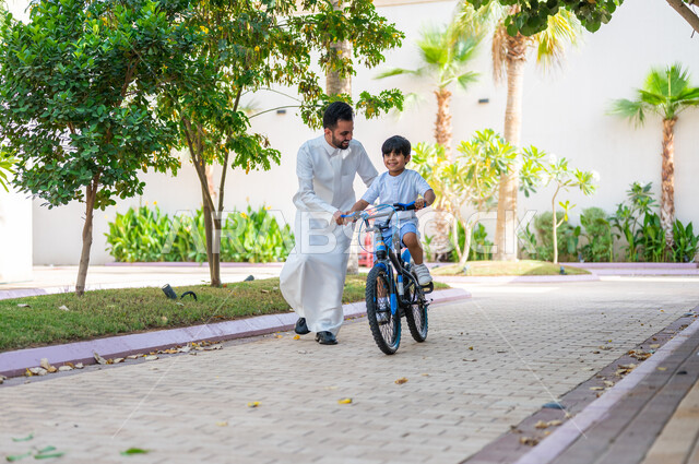 Gestures of joy in spending enjoyable family times, participating in recreational activities, helping children balance and playing on bicycles, a Saudi Gulf Arab father wearing traditional clothing teaching his son to ride a bike in a public park, the role of fathers in the lives of children