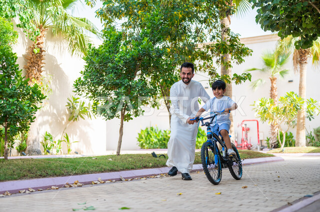 المشاركة في الأنشطة الترفيهية، مساعدة الأولاد على التوازن واللعب في الدراجات الهوائية، أب عربي خليجي سعودي يرتدي الزي التقليدي يعلم إبنه قيادة الدراجة في إحدى الحدائق العامة، إيماءات الفرح في قضاء أوقات عائلية ممتعة، دور الأباء في حياة الأبناء