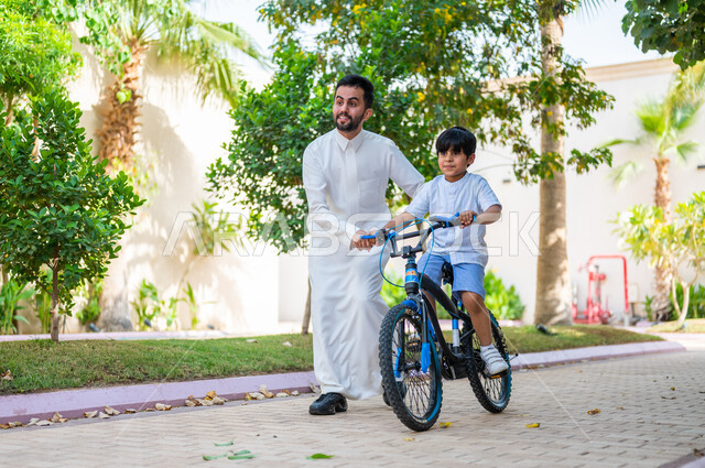 شاليهات ومنتجعات راقية في السعودية، ممارسة رياضة ركوب الدراجات الهوائيه, الاستمتاع بالاوقات عالائلية في وقت العطلة الصيفية, تعابير الحماس والسرور، أنشطة ترفيهيه مسلية, ولد عربي خليجي سعودي يقوم بقيادة الدراجة بمساعدة والده