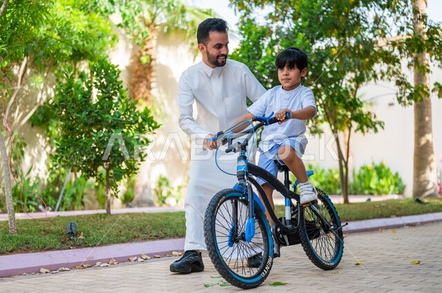 منتجعات وشاليهات راقية في السعودية، ممارسة رياضة ركوب الدراجات الهوائيه, الاستمتاع بالاوقات عالائلية في وقت العطلة الصيفية, تعابير الحماس والسرور، أنشطة ترفيهيه مسلية, ولد عربي خليجي سعودي يقوم بقيادة الدراجة بمساعدة والده