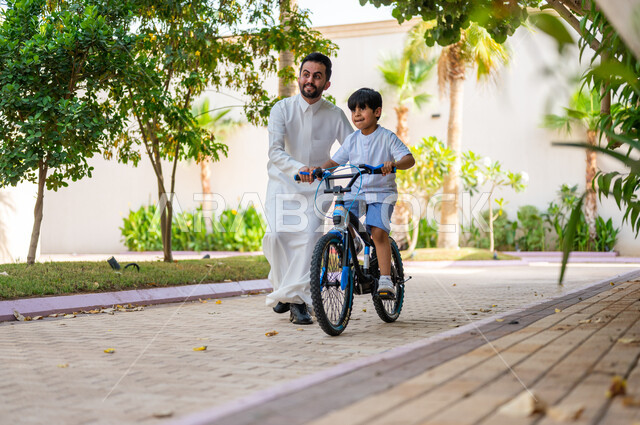 Practicing bicycle riding, enjoying family time during the summer vacation, expressions of enthusiasm and pleasure, entertaining recreational activities, a Saudi Gulf Arab boy riding a bicycle with the help of his father, upscale resorts and chalets in Saudi Arabia