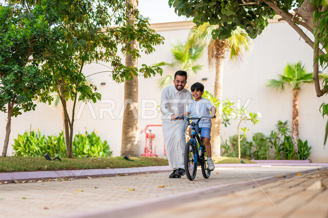 أنشطة ترفيهيه مسلية, ولد عربي خليجي سعودي يقوم بقيادة الدراجة بمساعدة والده, ايماءات الحماس والسرور، قضاء اوقات عائلية ممتعة في وقت العطلة الصيفية, ممارسة رياضة ركوب الدراجات الهوائيه, منتجعات وشاليهات راقية في السعودية