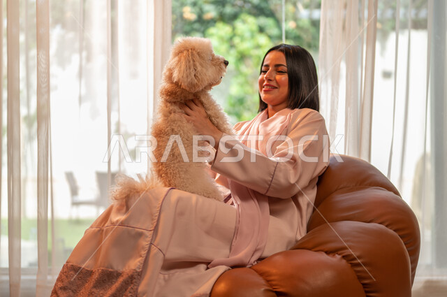 Expressions of pleasure and happiness, playing and having fun with pets, the concept of animal welfare, raising and owning domestic dogs, a smiling Saudi Arabian Gulf woman sitting on the sofa in the living room and holding a cute puppy in her hand.