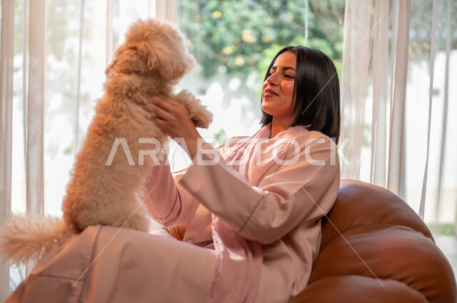 Playing and having fun with pets, the concept of animal welfare, raising and owning domestic dogs, a smiling Saudi Arabian Gulf woman sitting on the sofa in the living room and holding a cute puppy in her hand, gestures of pleasure and happiness