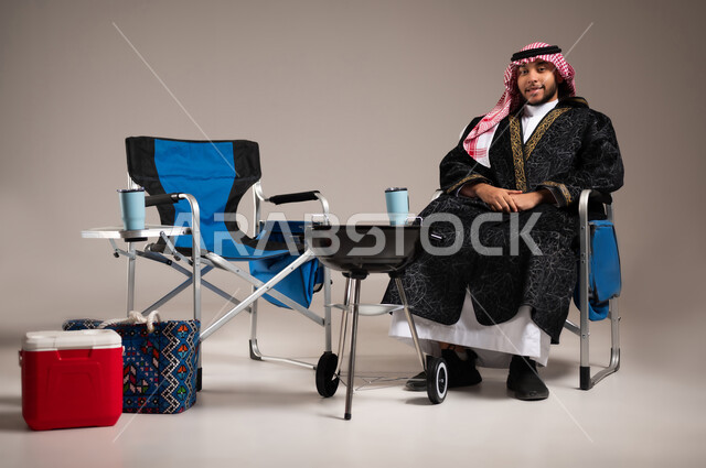 حفلة شواء في الإجازة , أنشطة ترفيهية ممتعة  , بورتريه لشاب عربي خليجي سعودي يرتدي الثوب التقليدي و الشماغ مع ملابس الشتاء الدافئة يجلس على الكرسي , النظر إلى الكاميرا بإيماءات الفرح و السرور , خلفية رمادية